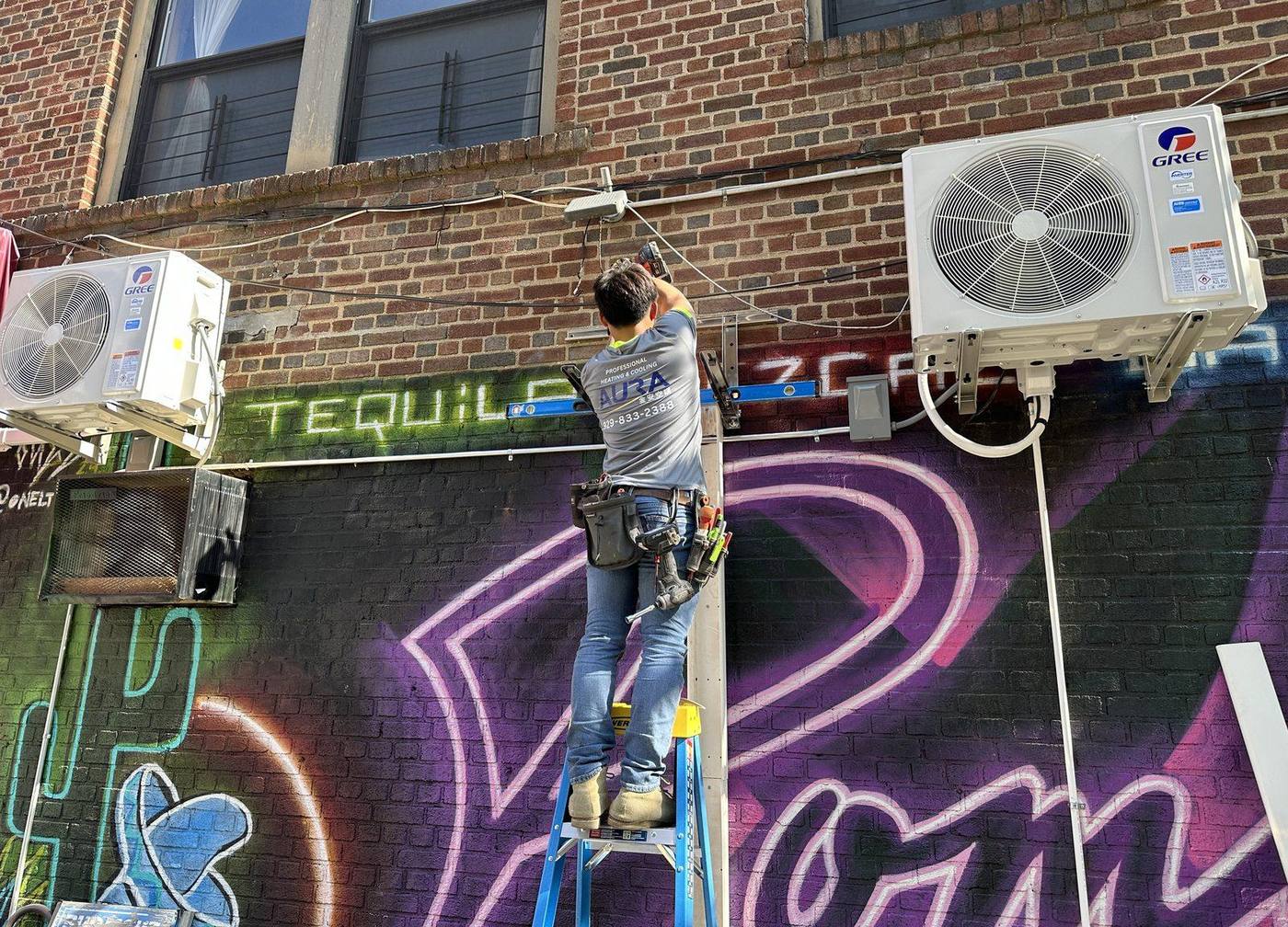 Aura HVAC technician on a ladder installing Gree mini-split outdoor units on a Brooklyn building